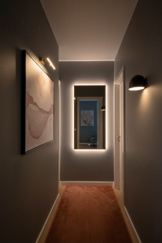 Couloir deco moderne avec éclairage ciblé : spot sur tableau abstrait, miroir à LED intégrée, applique d'ambiance et tapis velours rouille.