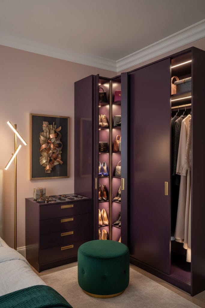 Dressing d'angle sur-mesure glamour en L laqué aubergine le soir. Portes à rotules centrales, niches à sacs éclairées, tiroirs à bijoux, compartiments à chaussures inclinés. Poignées laiton, éclairage LED dramatique et pouf vert émeraude. Ambiance luxueuse et intime.