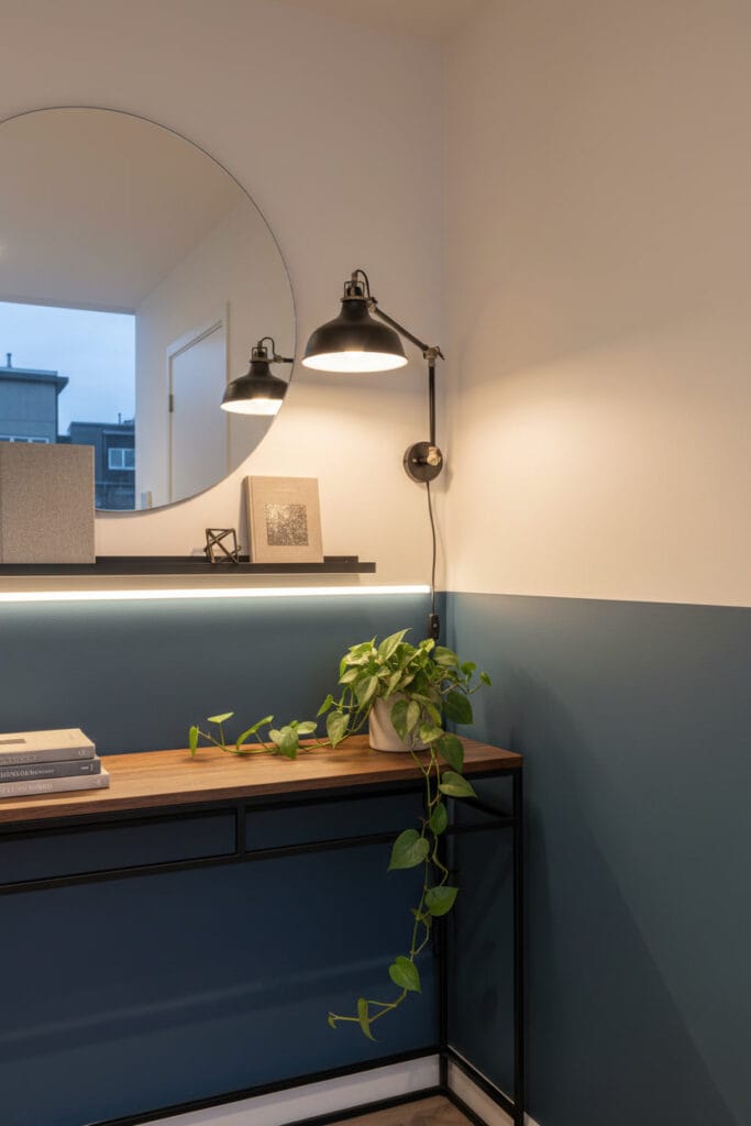 Coin de couloir industriel bicolore (blanc et bleu ardoise) avec bande LED intégrée, console murale métal, lampe industrielle, miroir sans cadre et étagère flottante, éclairé au crépuscule.